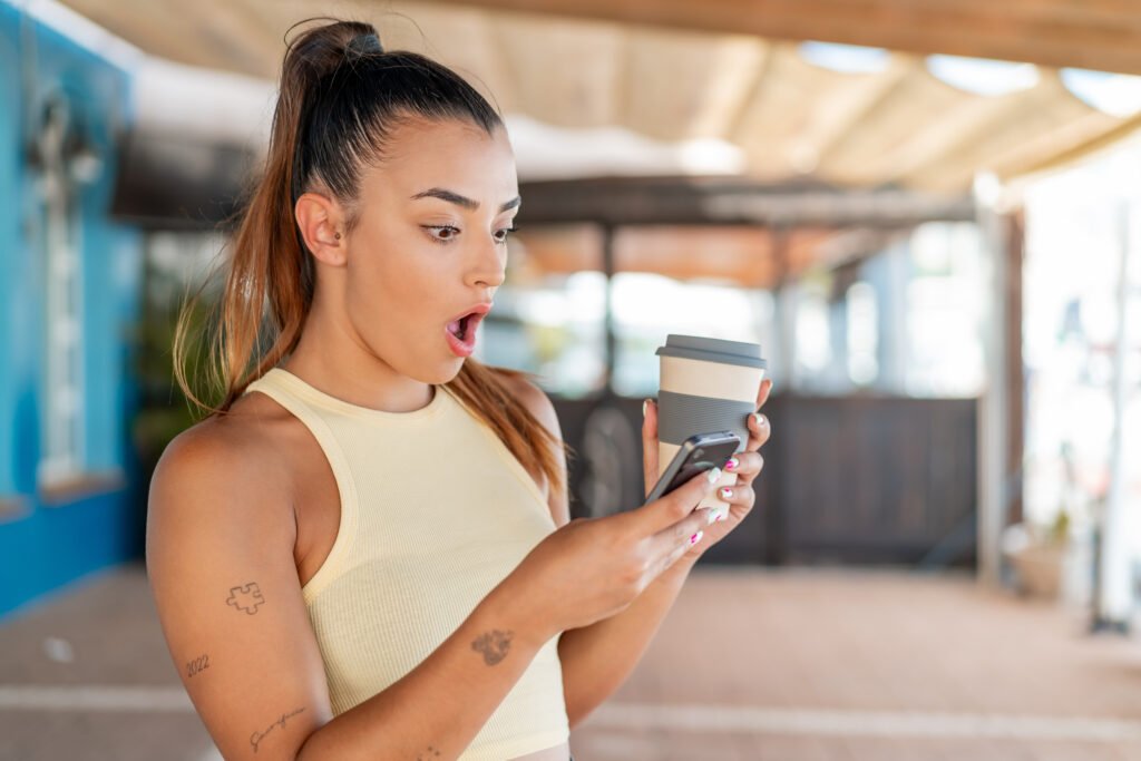 young pretty woman outdoors using mobile phone holding coffee with surprised expression
